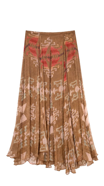 A pleated midi skirt in a brown base with abstract red, beige, and pink ikat-style patterns, shown flat on a light gray background.