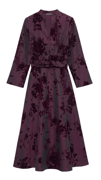 A long-sleeved wrap-style midi dress in a deep plum purple velvet fabric with a tonal floral jacquard pattern, featuring a matching waist tie belt, shown against a plain light gray background.