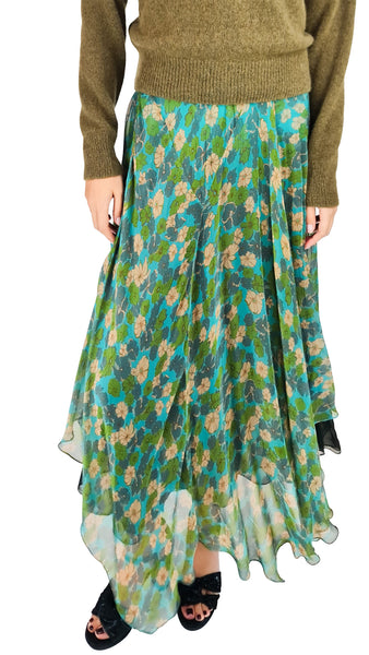 A model wearing a long, pleated high-low skirt with a green, teal, and beige floral pattern, paired with an olive green sweater and black sandals.