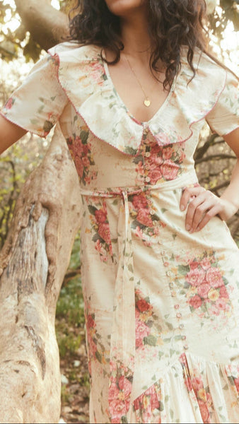 Woman in a floral dress standing in a garden with white flowers and sunlight filtering through.