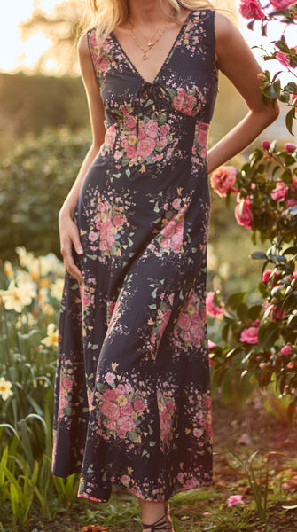 Woman in a floral dress standing in a garden with flowers and greenery.