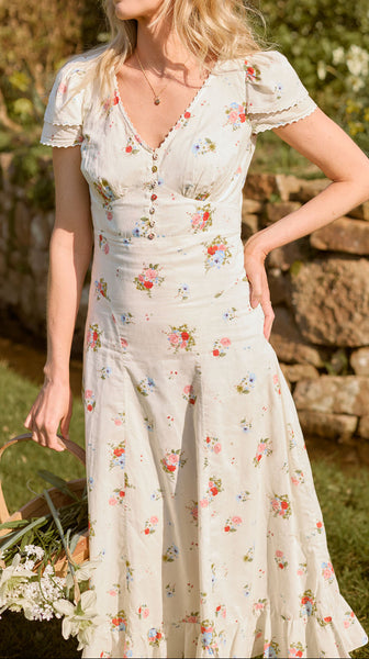 Woman in a floral dress standing outdoors with greenery and a stone wall in the background