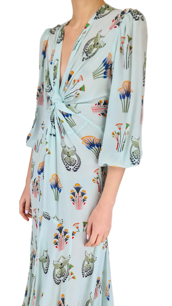 Woman wearing a long, light blue dress with colorful floral and bird pattern on a plain background