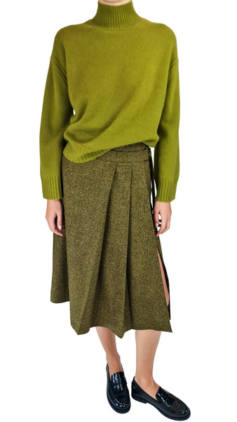 A model wearing a bright olive green turtleneck sweater and a textured olive green pleated midi skirt with side slits, paired with black leather loafers, against a plain white background.