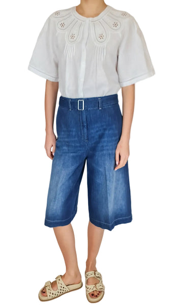 Person wearing a white blouse with floral details and blue denim shorts on a plain background
