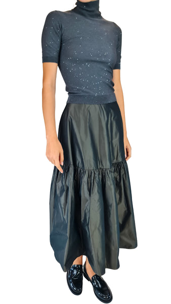 A model wearing a dark slate blue short-sleeve turtleneck top with subtle speckled details and a matching dark green tiered satin maxi skirt, paired with black patent leather loafers, against a plain white background.