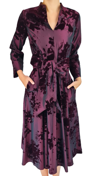 A woman wearing a long-sleeve wrap-style midi dress with a tie waist and pockets, featuring a dark purple and black floral jacquard pattern on a white background.