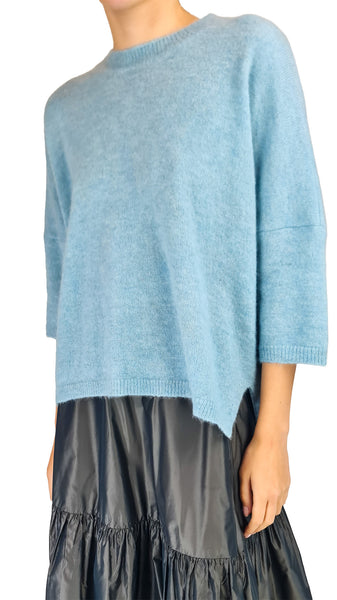 A person wearing a light blue knit sweater with three-quarter sleeves and an asymmetrical hem, paired with a dark pleated skirt, against a plain white background.