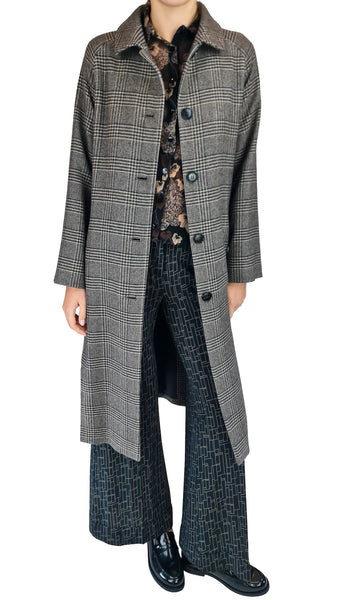 A model wearing a long gray and black plaid overcoat over a patterned top and dark wide-leg trousers, photographed against a plain white background.