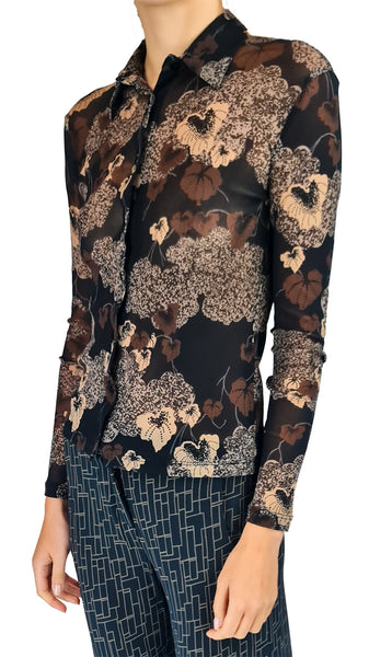 A woman wearing a sheer black long-sleeve button-up blouse with a brown and beige floral pattern, paired with dark patterned trousers, shown against a plain white background.