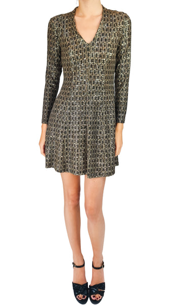 A model wearing a long-sleeve V-neck mini dress with a black and gold geometric lattice pattern, paired with black strappy high heels, against a plain white background.
