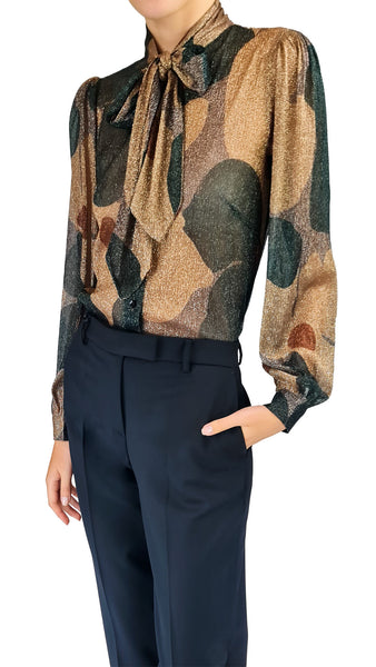 A woman wearing a sheer long-sleeve blouse with a tie-neck bow and an abstract organic pattern in beige, brown, and dark green, paired with dark navy trousers against a white background.