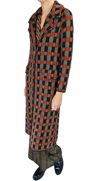 A model wearing a long-sleeved, collared coat with a bold orange, black, and gray checkered pattern, paired with patterned trousers and black leather shoes, shown against a plain white background.