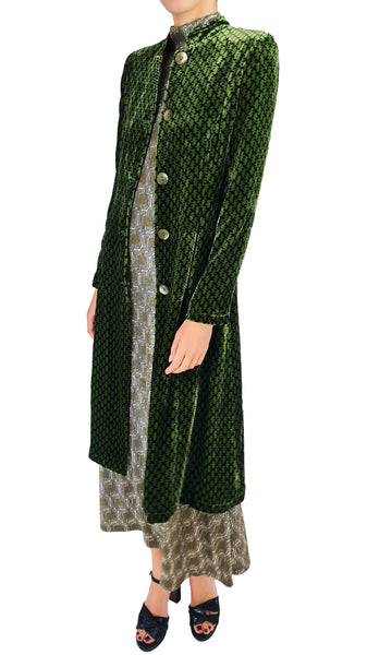 A model wearing a dark green patterned velvet long coat over a patterned dress, paired with black strappy heels, against a plain white background.