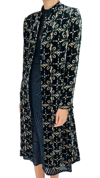 A long-sleeved open-front cardigan or coat in black velvet with a beige geometric and floral pattern, worn over a dark patterned dress. The image is cropped from the neck down against a plain white background.