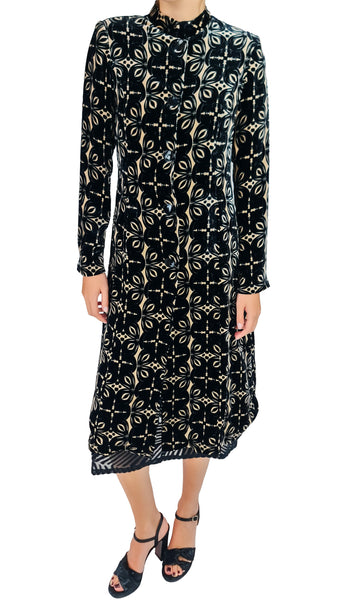 A long-sleeve midi dress with a black base and a beige geometric and floral pattern, modeled with black strappy heels against a white background.
